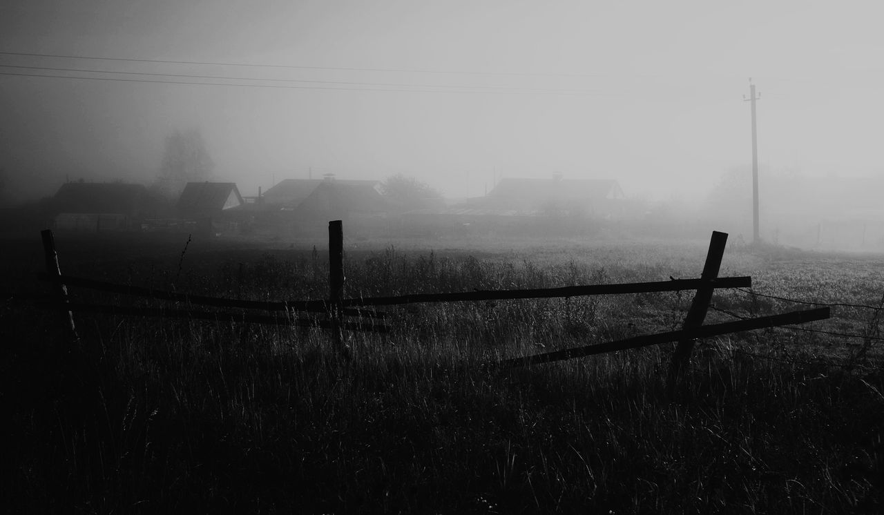 Mgła nad polem, drewniany płot, w tle zarysy zabudowań wsi o świcie